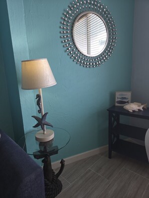 A room with teal walls, a round mirror with a metallic frame, a table with a lamp, and a framed picture of fish. - Oceanfront Paradise at Fountain Beach Resort / Daytona Beach (Daytona Beach)