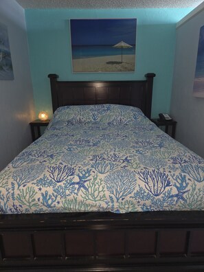 Un dormitorio con cama, mesita de noche, teléfono en la pared y un cuadro de una escena de playa.