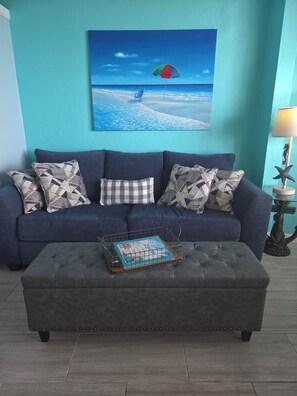 Ein Wohnzimmer mit einem blauen Sofa, einem grauen Hocker und einem gerahmten Bild mit einer Strandansicht.