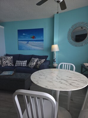 A living room with a blue wall, a ceiling fan, a round marble table, white chairs, a sofa with patterned cushions, a painting of a beach, and a decorative mirror. - Oceanfront Paradise at Fountain Beach Resort / Daytona Beach (Daytona Beach)