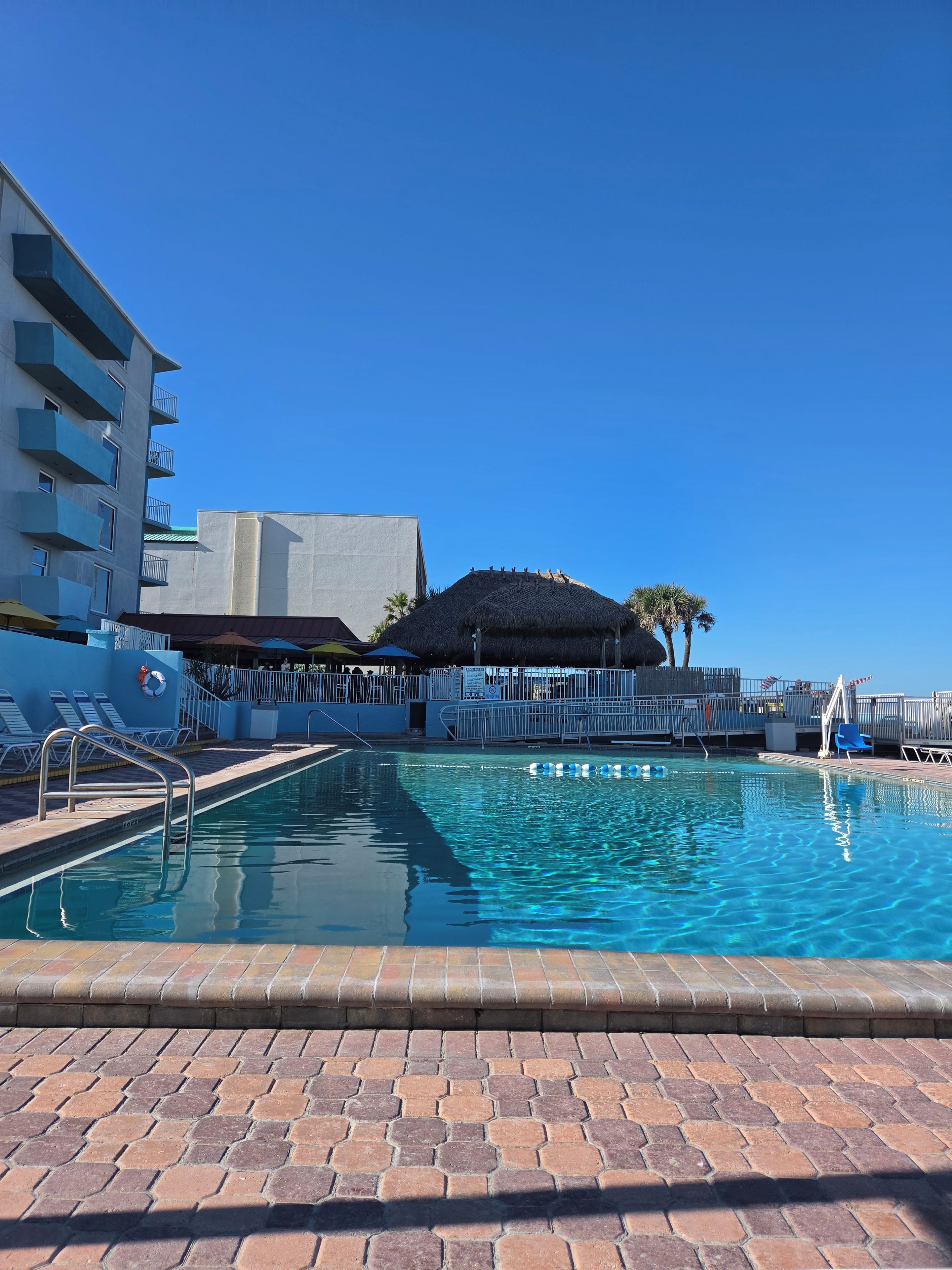Una piscina con borde de ladrillo, sillas de descanso y un edificio con balcones.