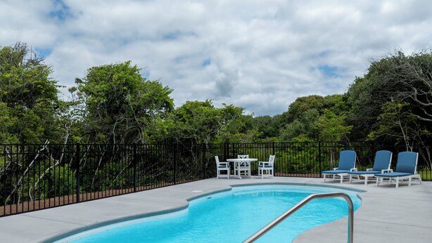 A swimming pool with a metal ladder, surrounded by a fence and trees, with lounge chairs and a table nearby. - Poolside retreat for large groups (North Topsail Beach)