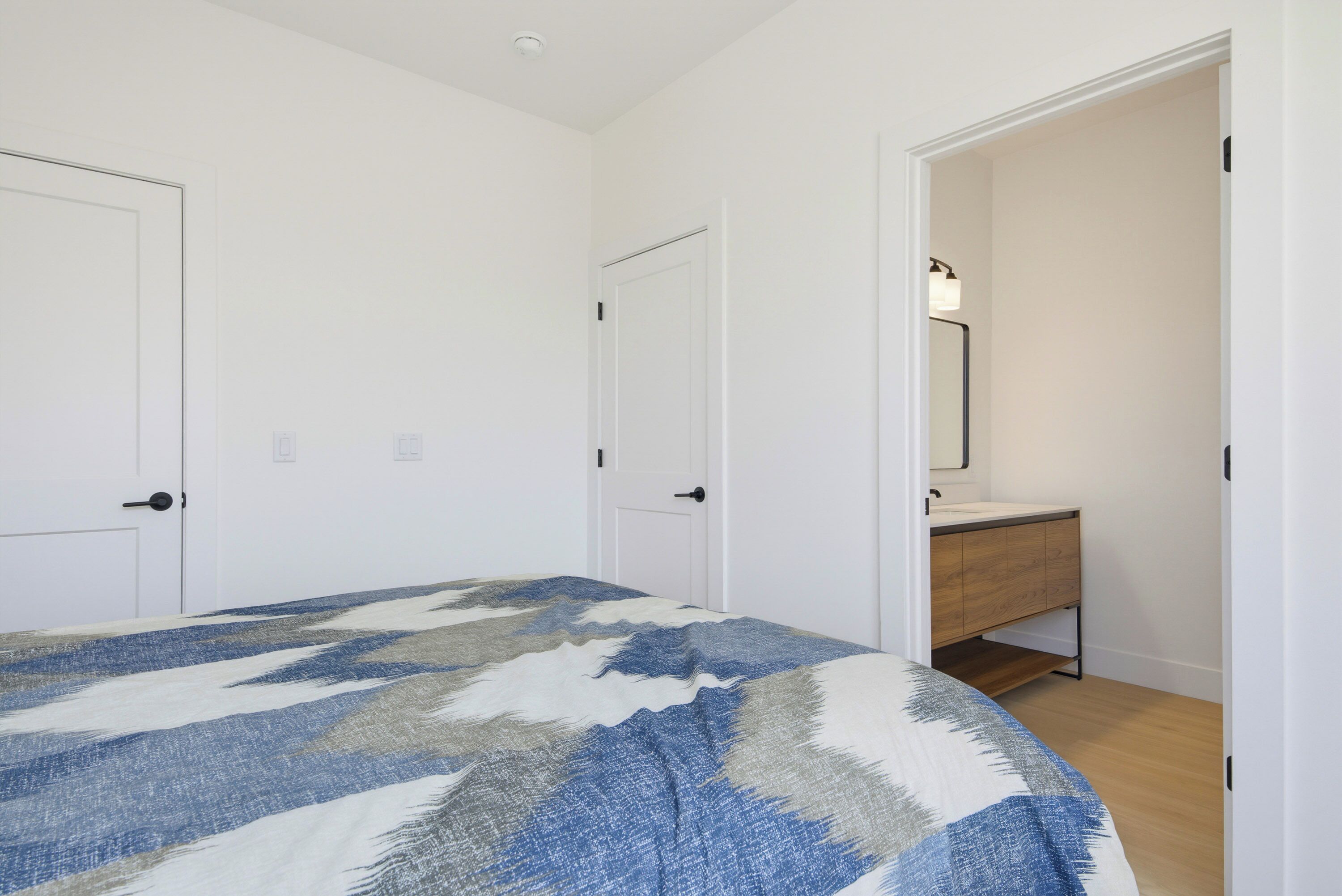 A bedroom with a bed, two doors, a bathroom with a mirror, and a wooden floor.