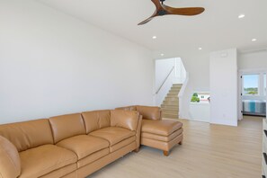 A modern living room with a tan leather sectional sofa, a wooden ceiling fan, and a staircase leading to an upper level. - Oceanfront pool retreat for large groups (North Topsail Beach)