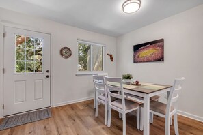 A dining area with a wooden table and white chairs, a window with a view of trees, and a wall-mounted picture. - Osceola's Oasis-Townhome with 3 bedrooms and 2.5 bathrooms. Walk to FSU/DOAK. (Tallahassee)