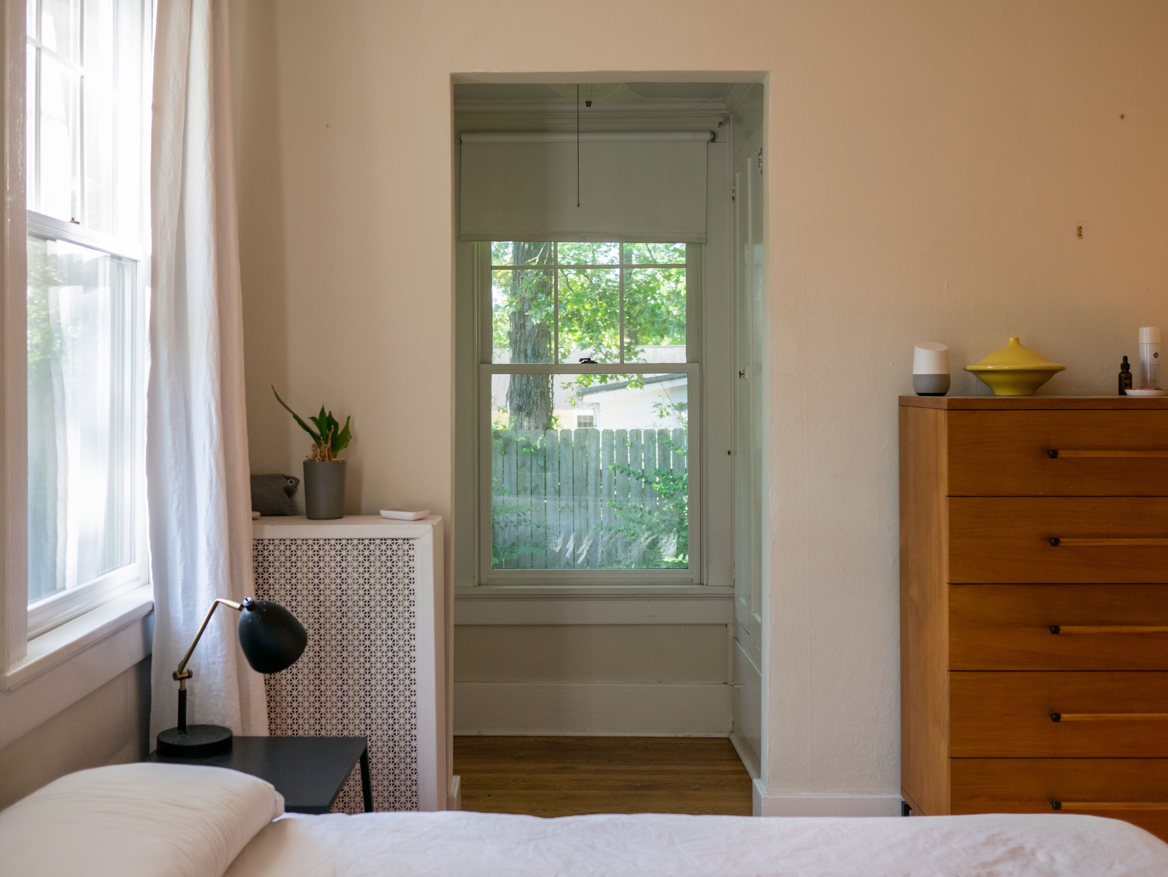 Une chambre à coucher comprenant un lit, une table de chevet avec une lampe, une commode, une fenêtre donnant sur une clôture et des arbres, ainsi qu’une petite table console blanche ornée d’une plante.