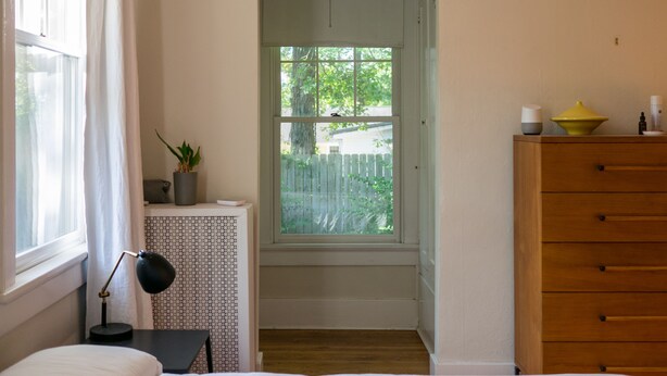 Ein Schlafzimmer mit Bett, Nachttisch mit Lampe, Kommode, Fenster mit Blick auf einen Zaun und Bäume sowie ein kleiner weißer Konsolentisch mit Pflanze.