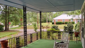 Eine überdachte Veranda mit grüner Kunstrasen, einem Schaukelstuhl und Blick auf eine Wohngegend mit einem Stoppschild und Bäumen.