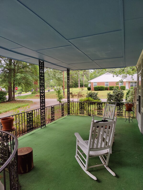 A covered porch with green artificial turf, a rocking chair, and a view of a residential area with a stop sign and trees. - Masters Rental: 3-bedroom house 1 block to Press Headquarters 1 mile to course (Augusta)