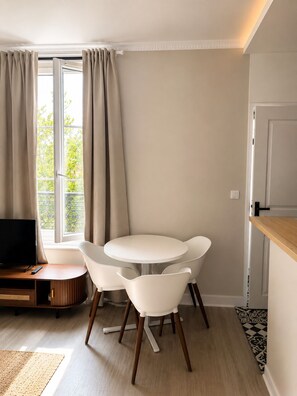 Un salon moderne avec une table ronde et deux chaises blanches, un téléviseur à écran plat et une fenêtre avec des rideaux.
