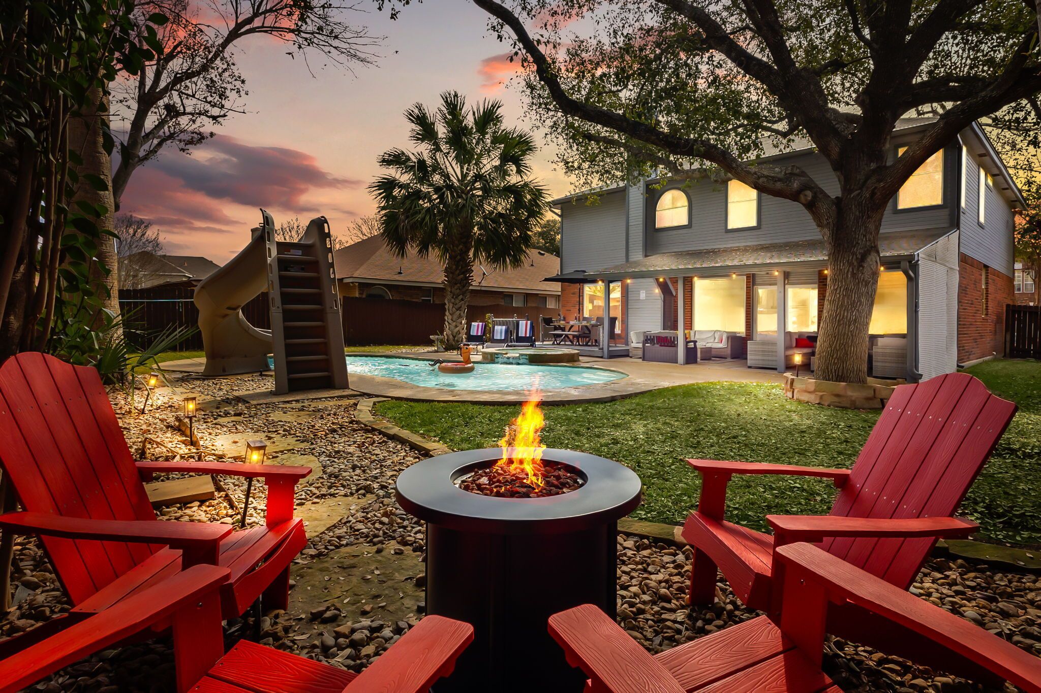 A backyard with a fire pit, red Adirondack chairs, a pool, and a house with a large tree.