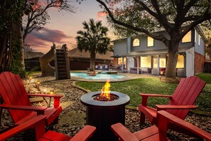 A backyard with a fire pit, red Adirondack chairs, a pool, and a house with a large tree. - Family Fun, Heated Pool, Slide, Hot Tub, Game Room (San Antonio)