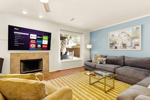 A living room with a flat-screen TV displaying streaming services, a fireplace, a sofa, a coffee table, and a painting on the wall. - Family Fun, Heated Pool, Slide, Hot Tub, Game Room (San Antonio)