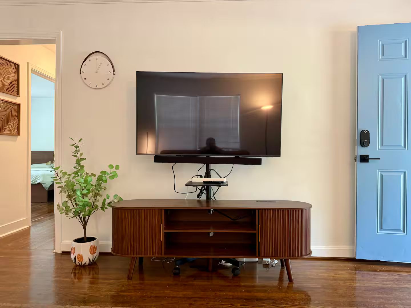 A living room with a flat-screen TV mounted on the wall, a wooden entertainment center, a potted plant, and a door with a keypad lock.