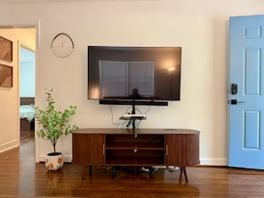 A living room with a flat-screen TV mounted on the wall, a wooden entertainment center, a potted plant, and a door with a keypad lock. - Serene 2BR + Study, Min to NoDa & Uptown (Charlotte)
