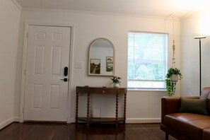 A room with a door, a console table with a mirror, a window with blinds, a brown leather chair, and a hanging plant. - Serene 2BR + Study, Min to NoDa & Uptown (Charlotte)