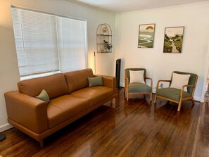 Ein Wohnzimmer mit braunem Ledersofa, zwei grünen Sesseln, Holzböden, einer Stehlampe und zwei an der Wand befestigten Bildern.