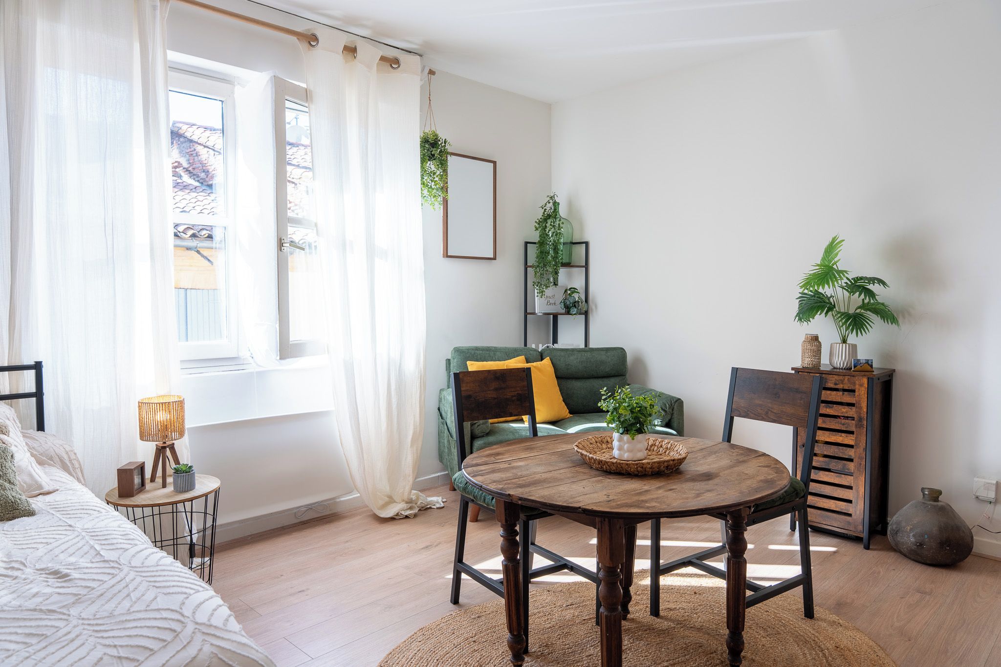 A well-lit room with a bed, a green sofa, a wooden table with a basket, and a chair. There is a window with curtains, a plant on a shelf, and a lamp on a side table.