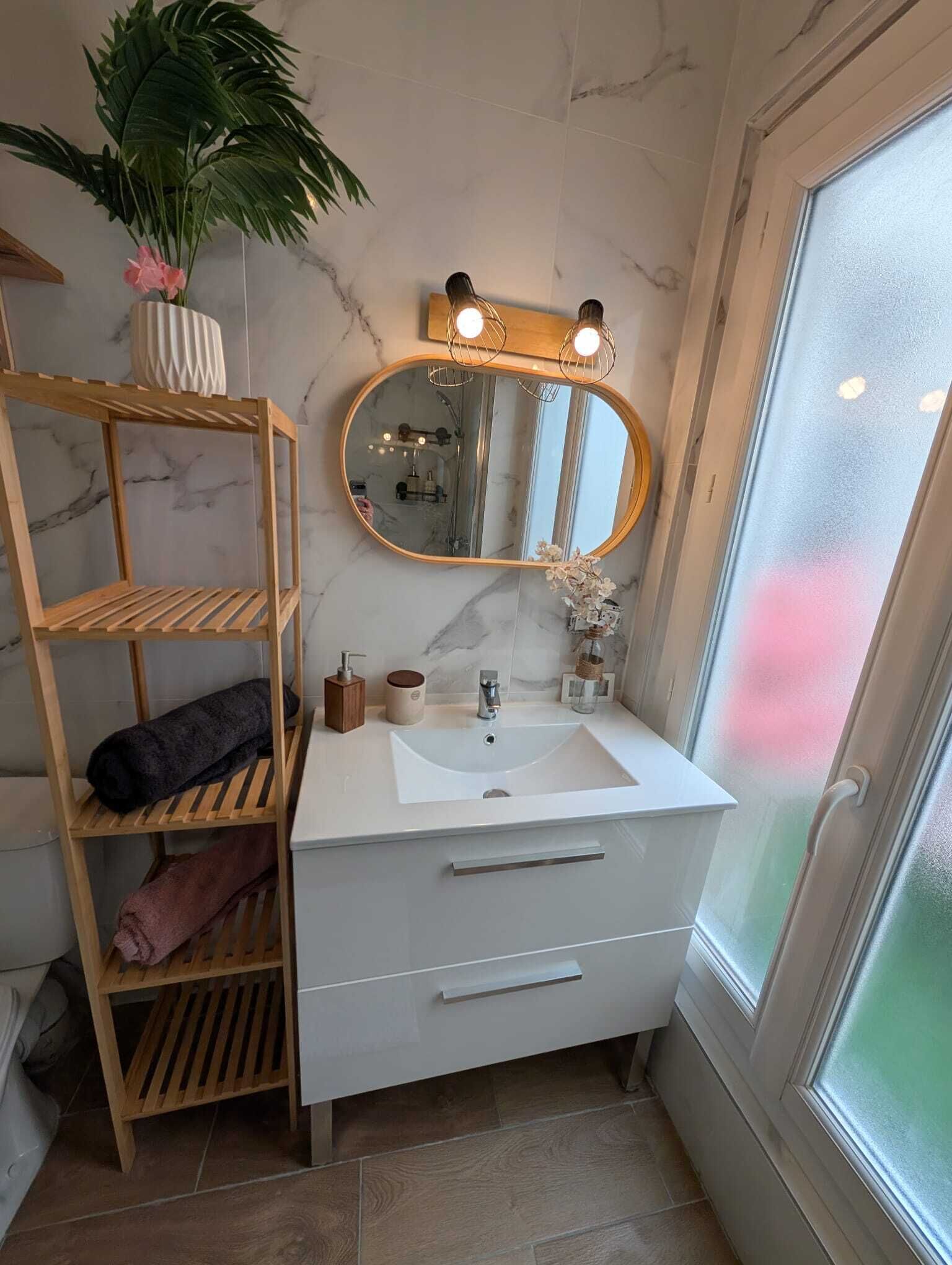 A bathroom with a white vanity, a mirror with lights, a wooden shelf, and a potted plant.