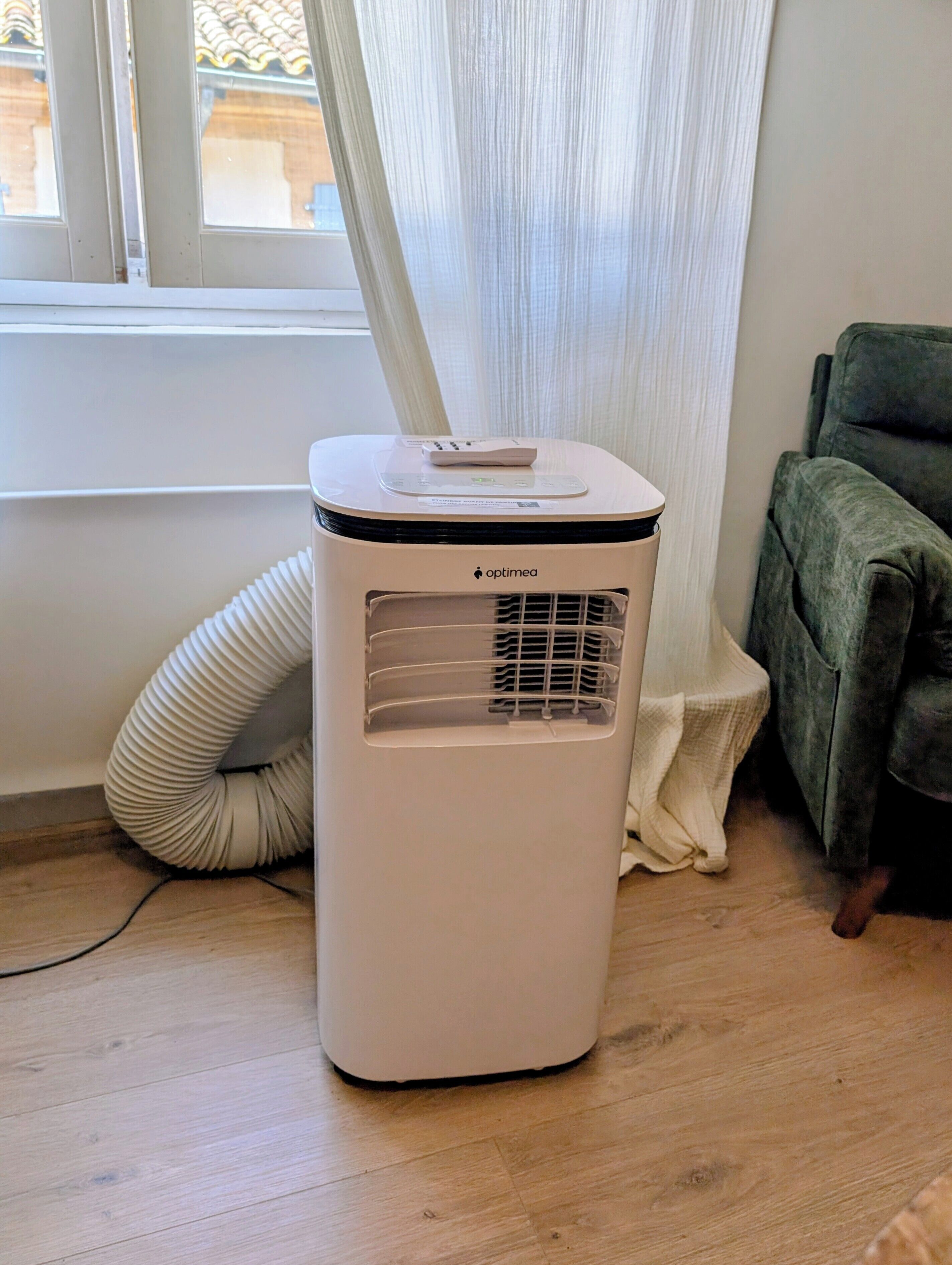 A white portable air conditioner with a control panel, placed on a wooden floor next to a window with white curtains.