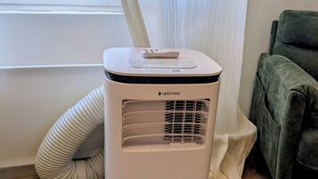 A white portable air conditioner with a control panel, placed on a wooden floor next to a window with white curtains.