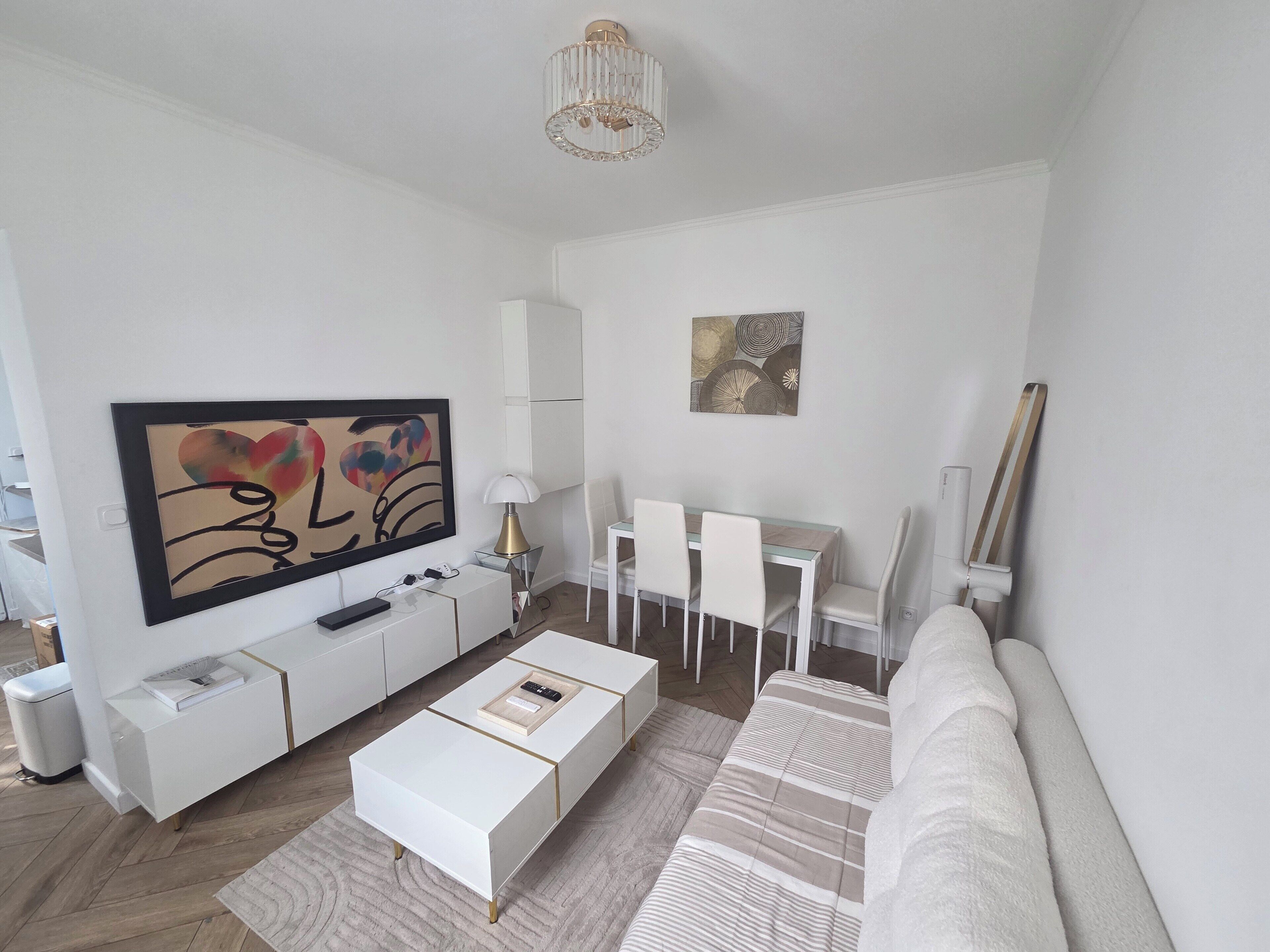 Ein modernes Wohnzimmer mit Flachbildfernseher, weißen Möbeln, einem Essbereich mit Stühlen und einer Couch.