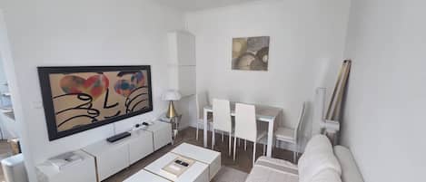 A modern living room with a flat-screen TV, white furniture, a dining area with chairs, and a sofa.