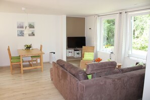 Een woonkamer met een bruine bank, een houten eettafel met stoelen, een televisie en een raam met gordijnen.