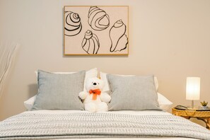 Siististi pedattu sänky, jossa on valko-harmaakuvioinen päiväpeite, valkoinen teddykarhu, jonka kaulassa on oranssi rusetti, ja harmaat tyynyt. Sängyn yläpuolella näkyy seinätaide, jossa on abstrakteja muotoja.