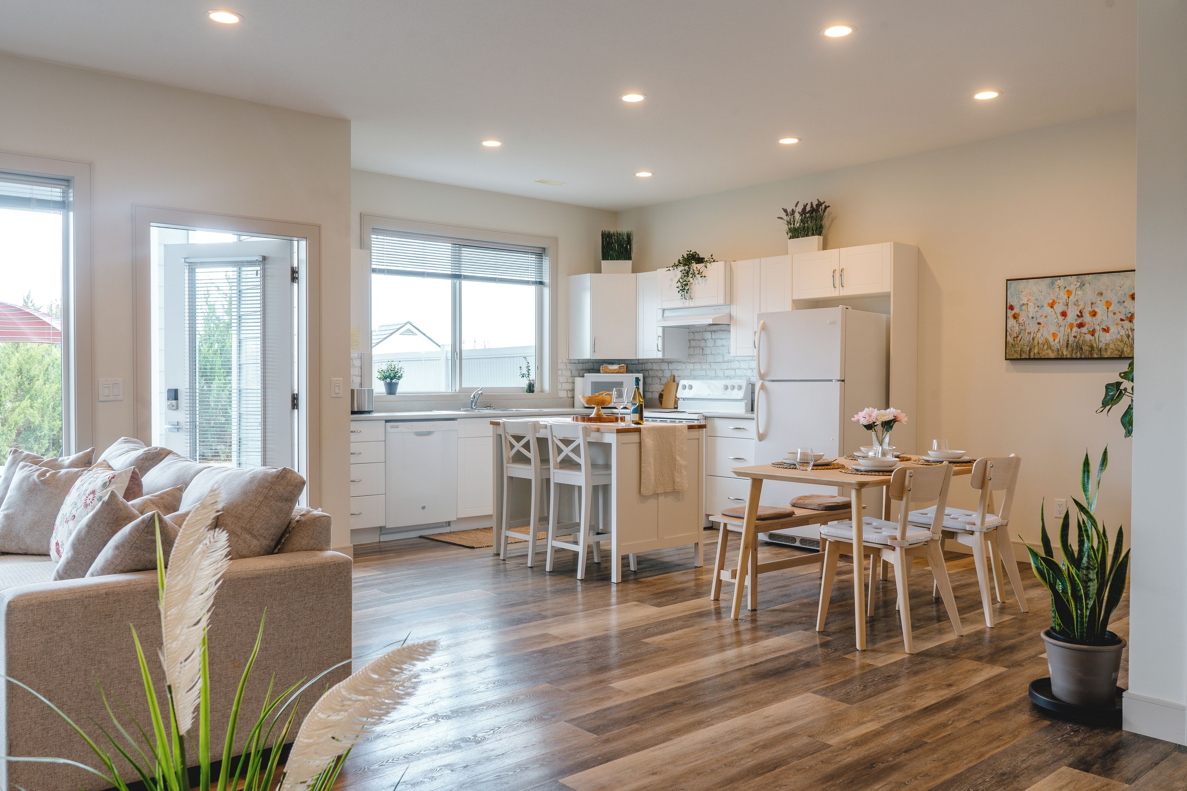 Una cocina moderna con armarios blancos, un comedor con mesa y sillas de madera, y una sala de estar con sofá color beige.