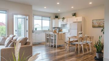 Una cocina moderna con armarios blancos, un comedor con mesa y sillas de madera, y una sala de estar con sofá color beige.