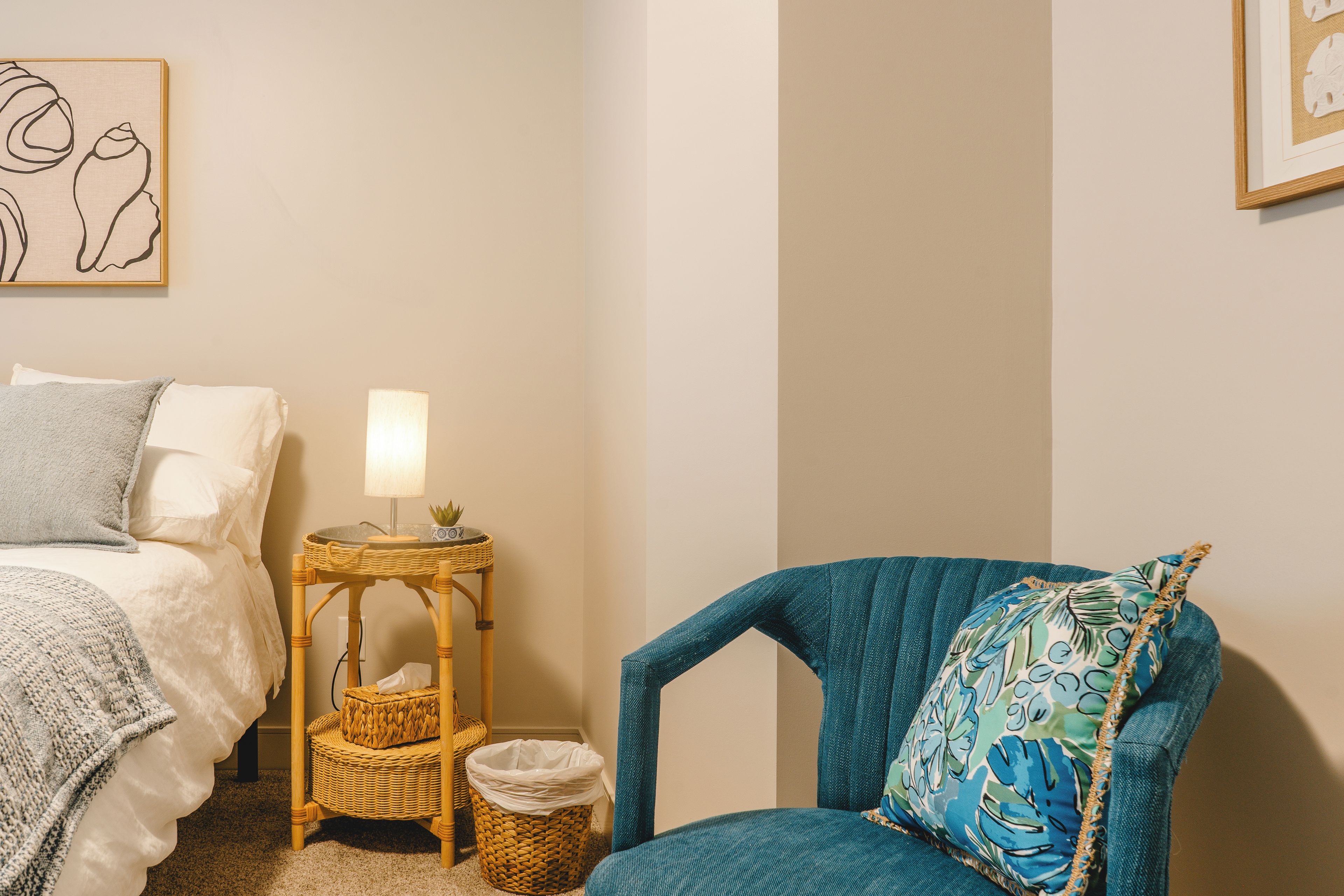Un dormitorio con una cama, una silla, una mesita de noche con una lámpara, una obra de arte en la pared y una cesta de mimbre.