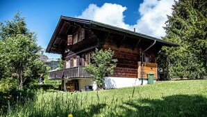 A traditional wooden chalet with a balcony, surrounded by lush greenery and a clear sky.