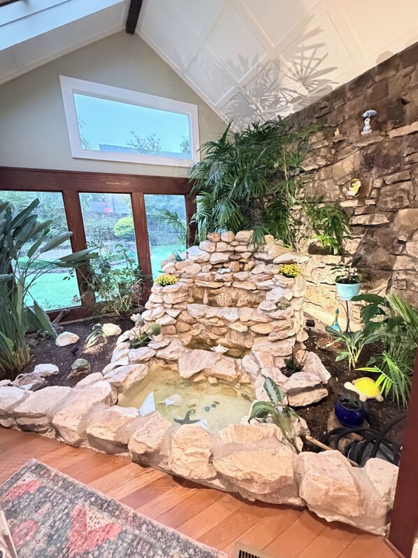 Jardín rocoso interior con fuente de agua, rodeado de diversas plantas y vistas al exterior a través de amplios ventanales.