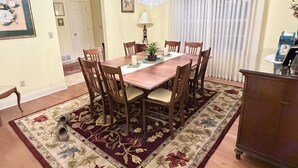 Een eetkamer met een houten tafel, zes stoelen, een kroonluchter, een tapijt met bloemenmotief en een raam met gordijnen.