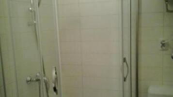 Zona de ducha con paredes de cristal, pared revestida de azulejos blancos y un inodoro blanco a la derecha.