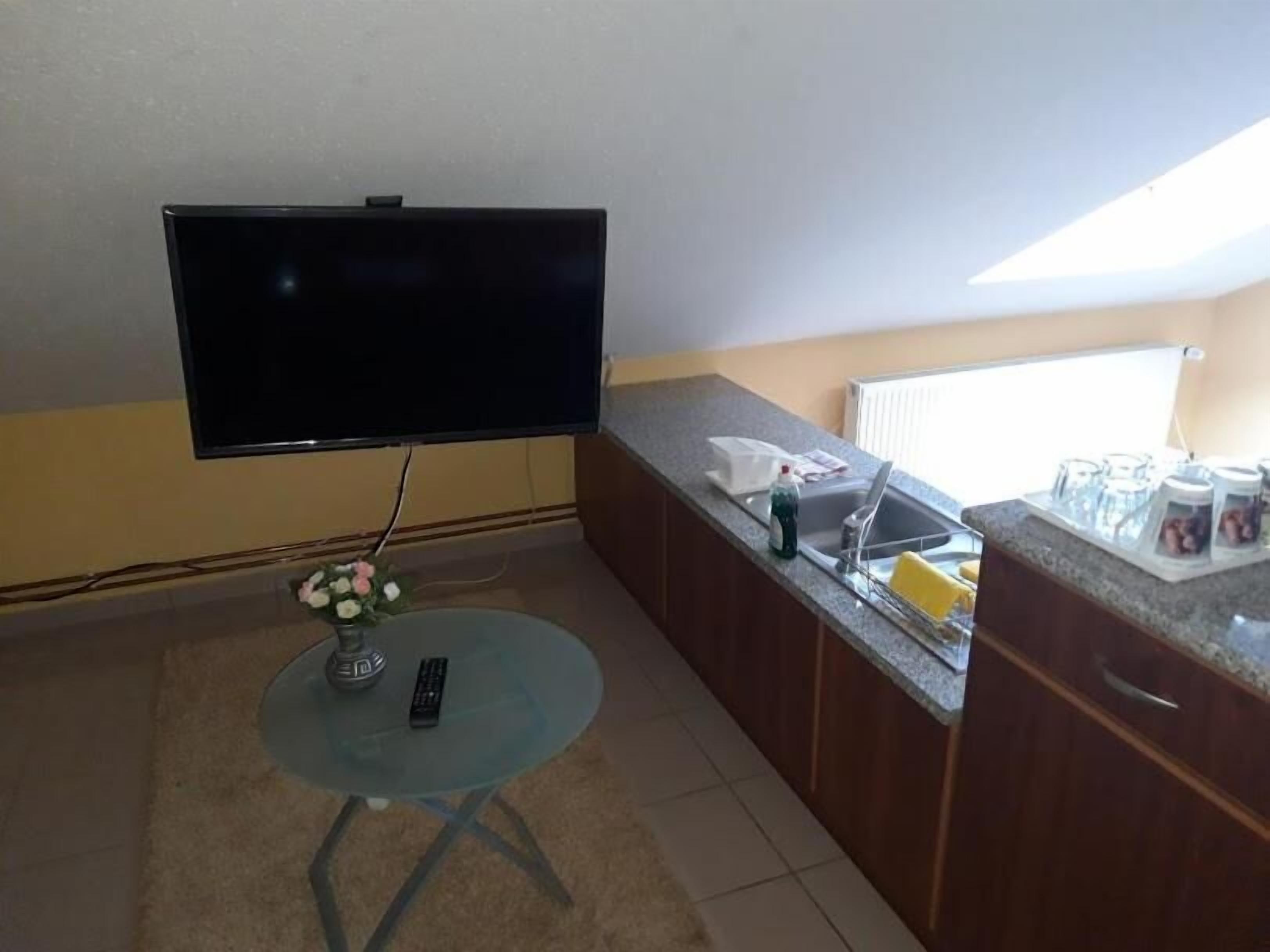 A kitchen with a flat-screen TV mounted on the wall, a small round table with a vase and remote control, and a countertop with kitchen items.
