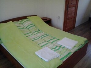 Una cama individual con una colcha verde y blanca a rayas, dos toallas blancas dobladas y una mesita de noche de madera con una pequeña mesa junto a ella.