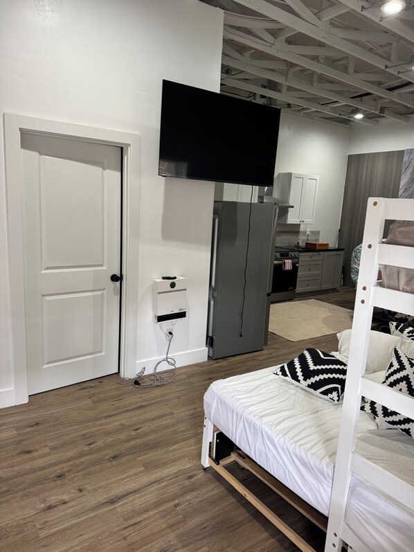 A modern bedroom with a bed, a TV mounted on the wall, and a kitchenette in the background.