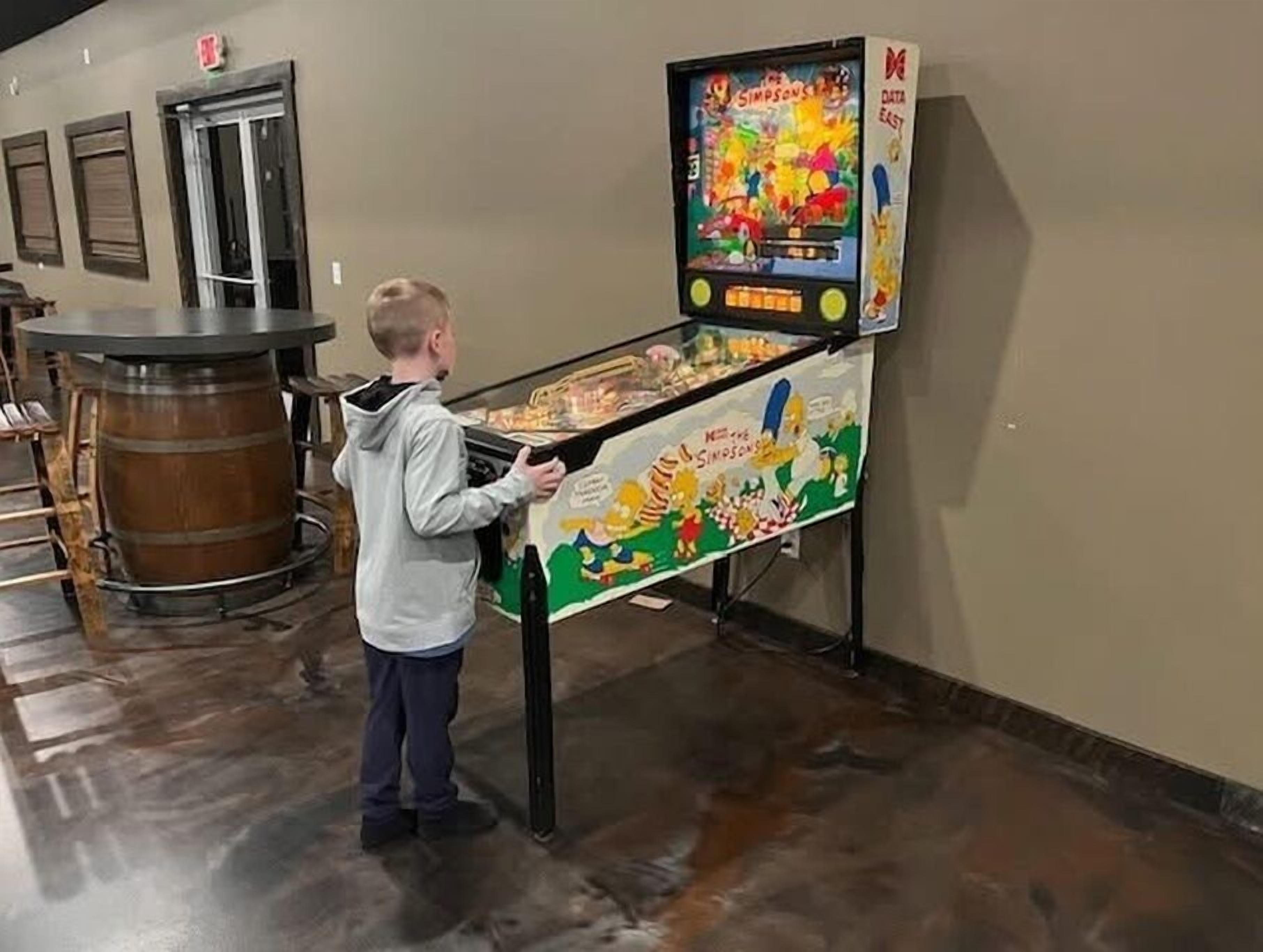 A child playing a pinball machine in a room with a wooden barrel and a door in the background.