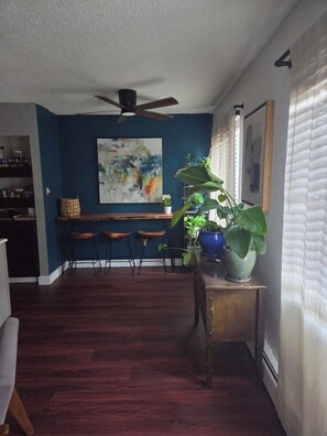 Ein Zimmer mit blauer Wand, Deckenventilator, Essbereich mit Hocker und einer Topfpflanze.