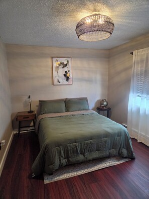 Ein Schlafzimmer mit Bett, Nachttisch, Lampe, Wandbild und einem Fenster mit Vorhängen.