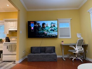 A living room with a flat-screen TV mounted on the wall displaying a movie poster, a gray sofa, a white desk with a chair, and a window with blinds. - A Cozy Stay on Coronation Street (St. John's)