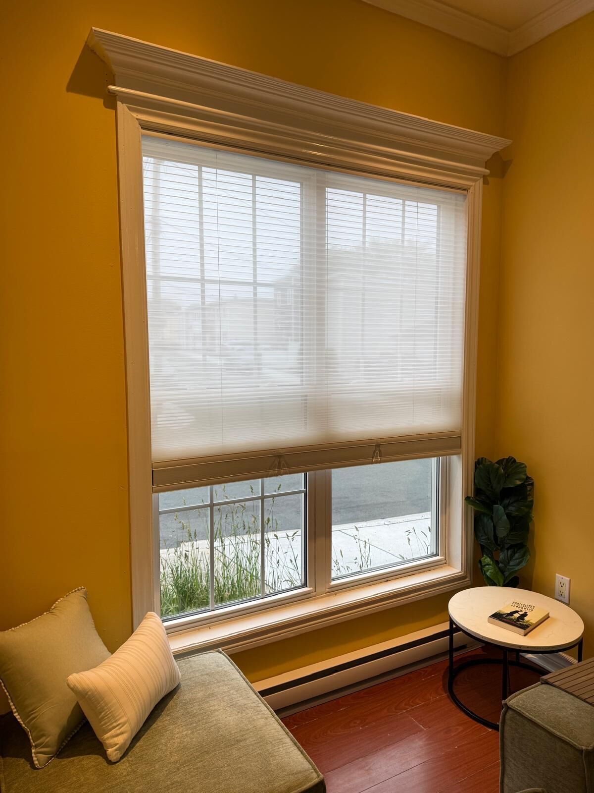 A room with a large window, a small round table with a book, a couch with cushions, and a potted plant.