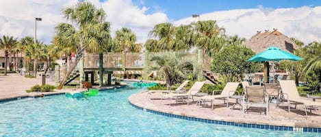 A swimming pool with lounge chairs and umbrellas, surrounded by palm trees and a thatched-roof structure.