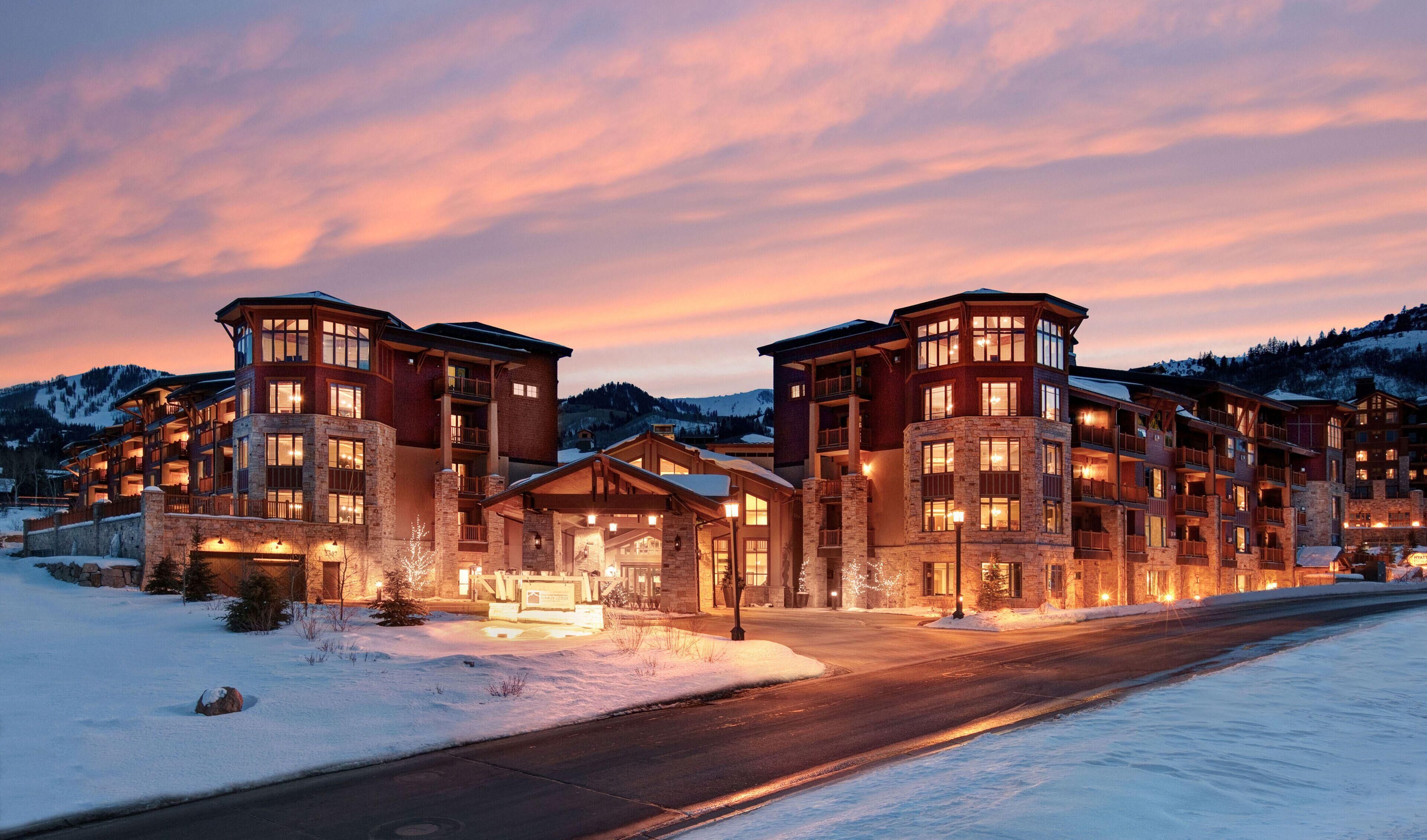 A multi-story hotel with a stone facade, large windows, and a snow-covered landscape at dusk.