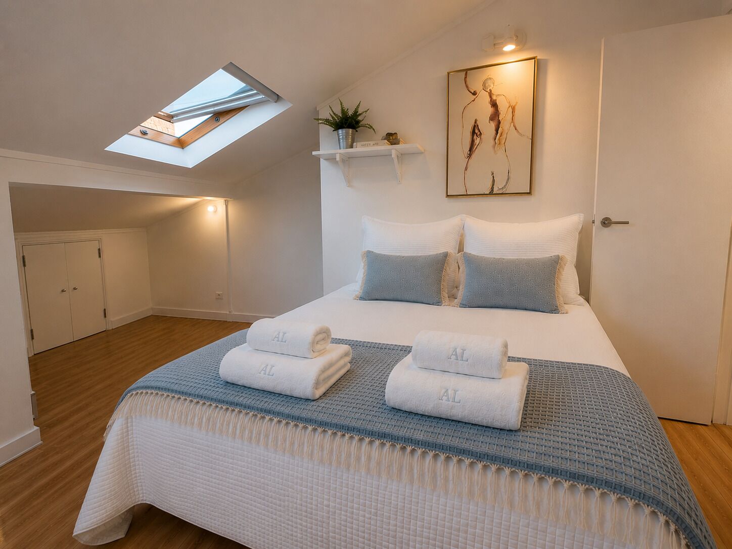 Une chambre avec un puits de lumière, un lit avec des serviettes, une étagère murale avec une plante et un tableau.