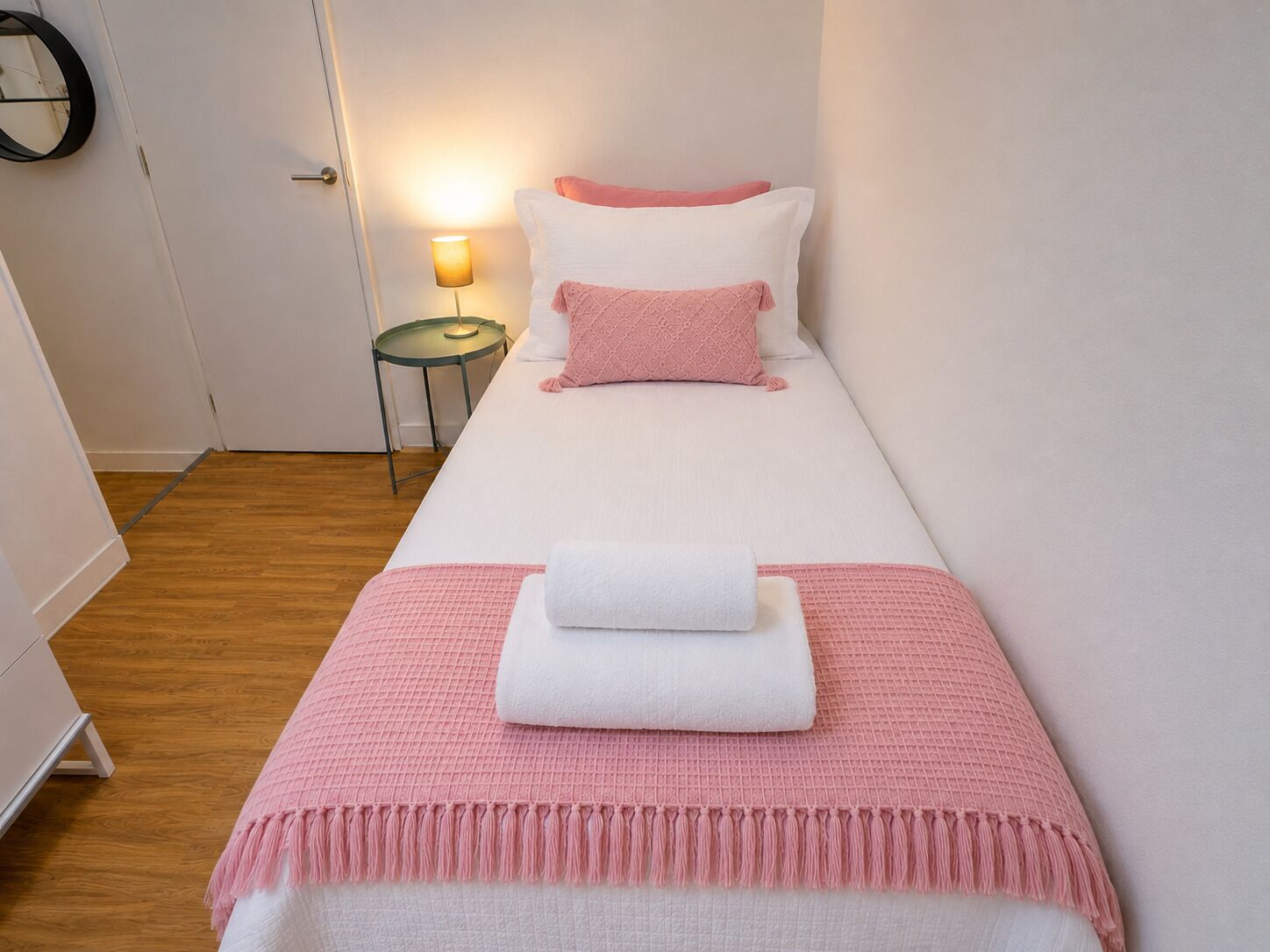Ein Einzelbett mit einem rosa-weißen Bettwäsche-Set, ein kleiner Nachttisch mit Lampe und ein runder Spiegel an der Wand.