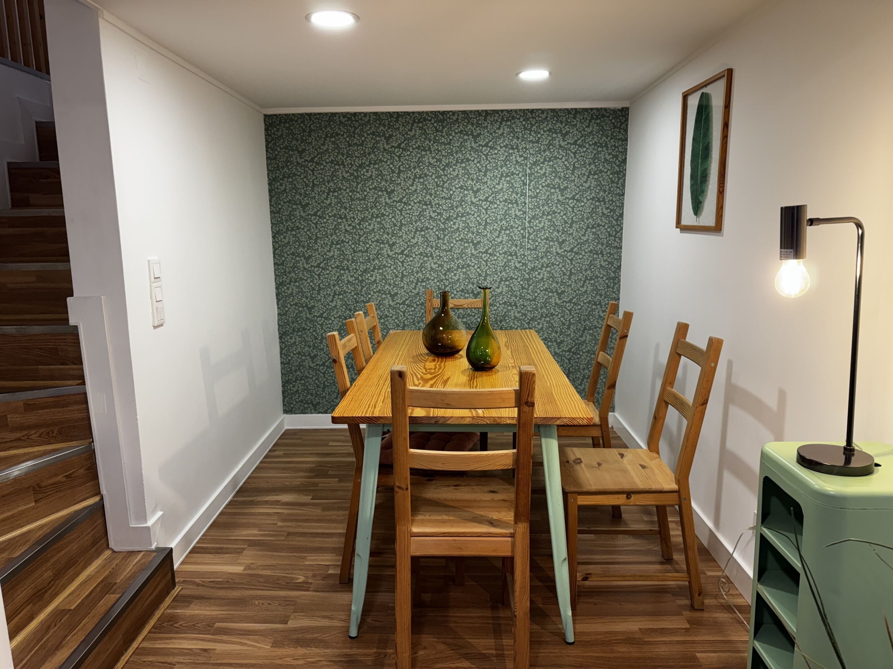 Ein Essbereich mit einem Holztisch und Stühlen, einer Wand mit grünem Muster, einem gerahmten Bild und einer Stehlampe.
