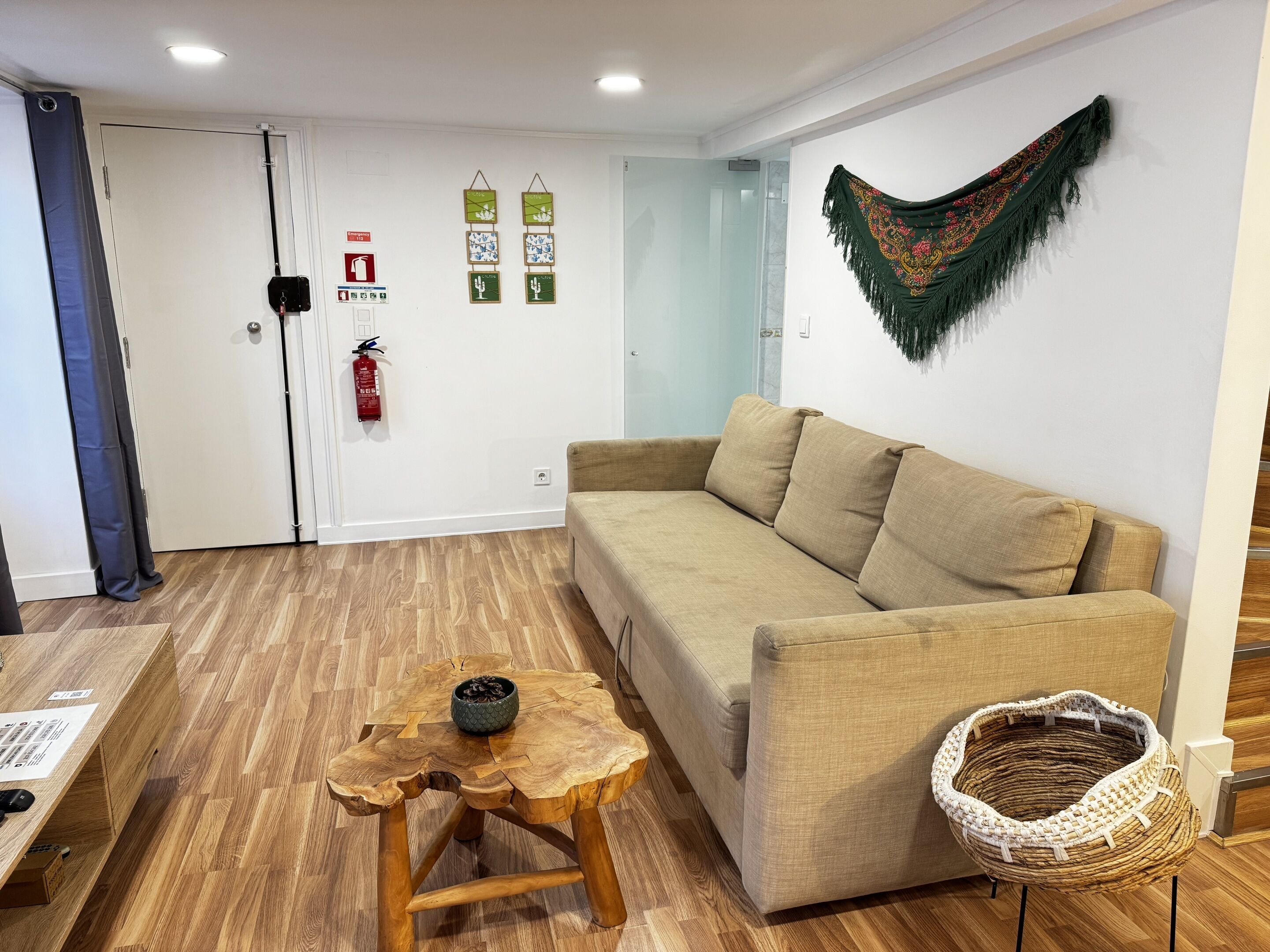 A room with a beige sofa, a woven chair, a wooden coffee table, and a fire extinguisher on the wall.